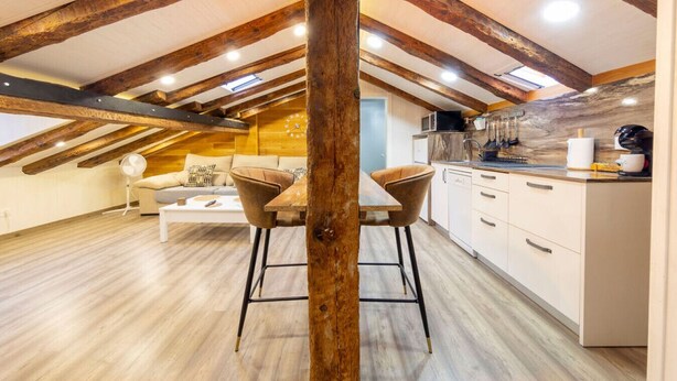 Un apartamento moderno estilo loft con vigas de madera a la vista, una cocina con armarios blancos y un comedor con una mesa de madera y dos sillas.