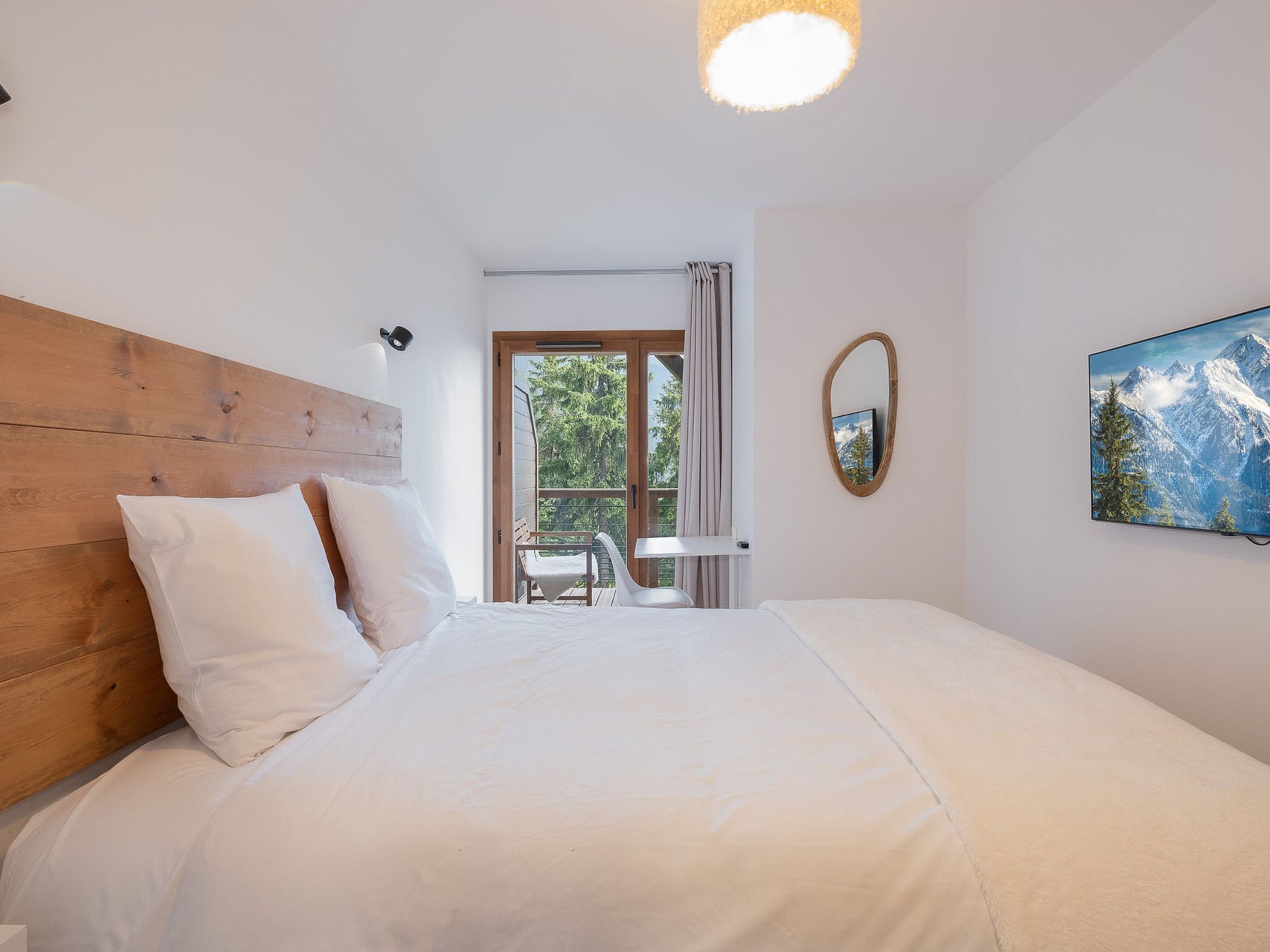 A bedroom with a wooden headboard, a bed with white bedding, a window with curtains, a chair, and a painting on the wall.