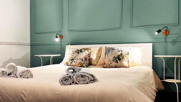 A neatly made bed with floral pillows and rolled towels, a white bedside table, and a wall-mounted lamp. - Raffy in Rome (Rome)
