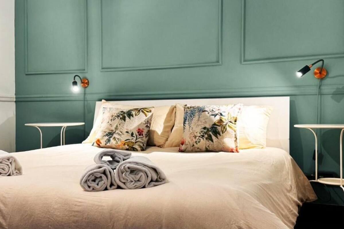A neatly made bed with floral pillows and rolled towels, a white bedside table, and a wall-mounted lamp.