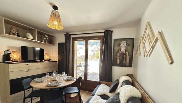 A cozy living room with a sofa, a dining table set for four, a TV, and a large window with curtains. - Cozy 2-room apartment, terrace, ski locker, shared parking (Demi-Quartier)