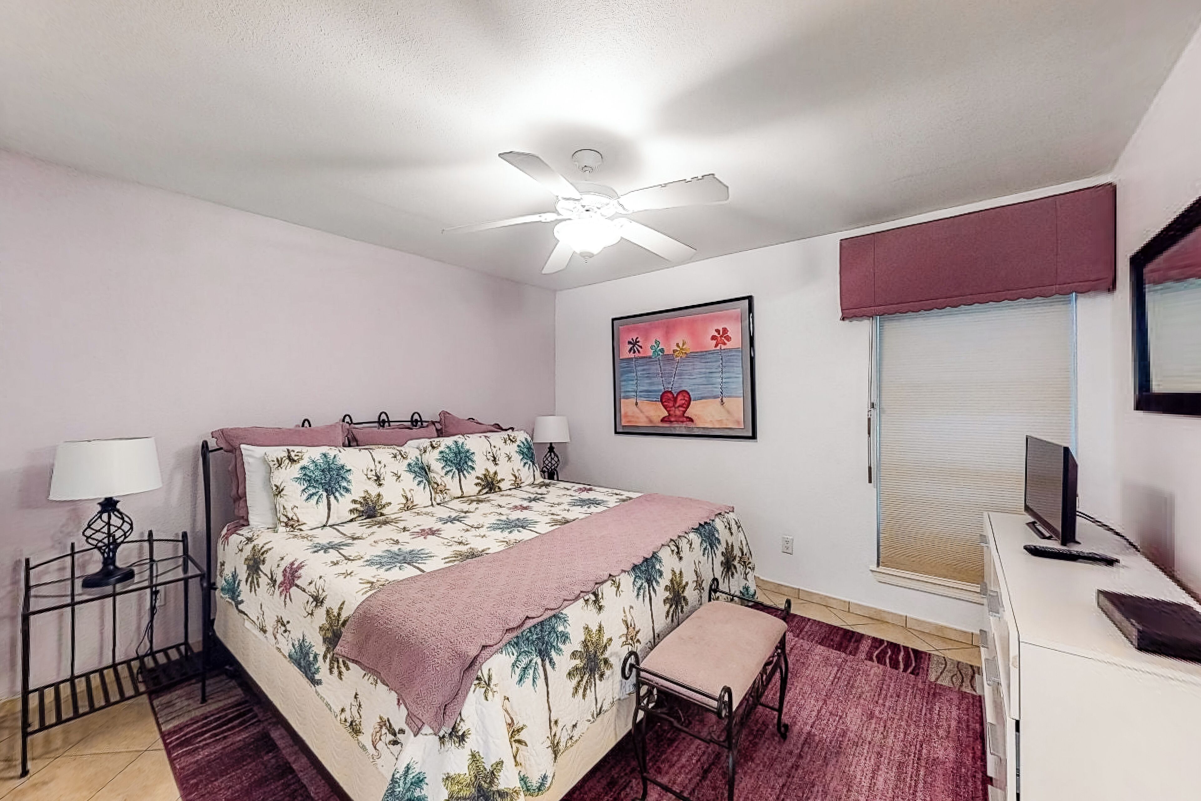 A bedroom with a bed, a desk, a chair, a ceiling fan, a window with blinds, and a wall-mounted TV.