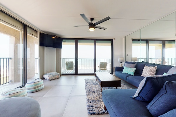 A modern living room with a blue sofa, a coffee table, and a striped rug. There is a ceiling fan and a TV mounted on the wall. - 3BR w/ Wraparound Balcony: Suntide III - Unit 901 (South Padre Island)