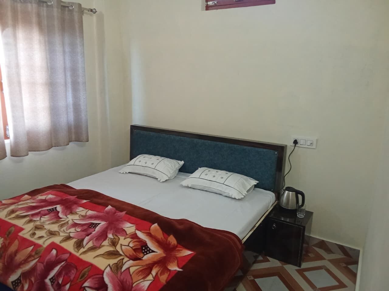 A neatly made bed with a floral bedspread, a bedside table with a kettle, and a power outlet on the wall.