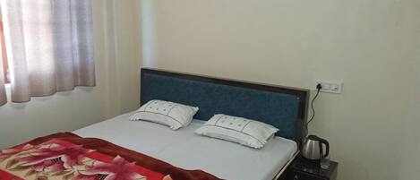 A neatly made bed with a floral bedspread, a bedside table with a kettle, and a power outlet on the wall.