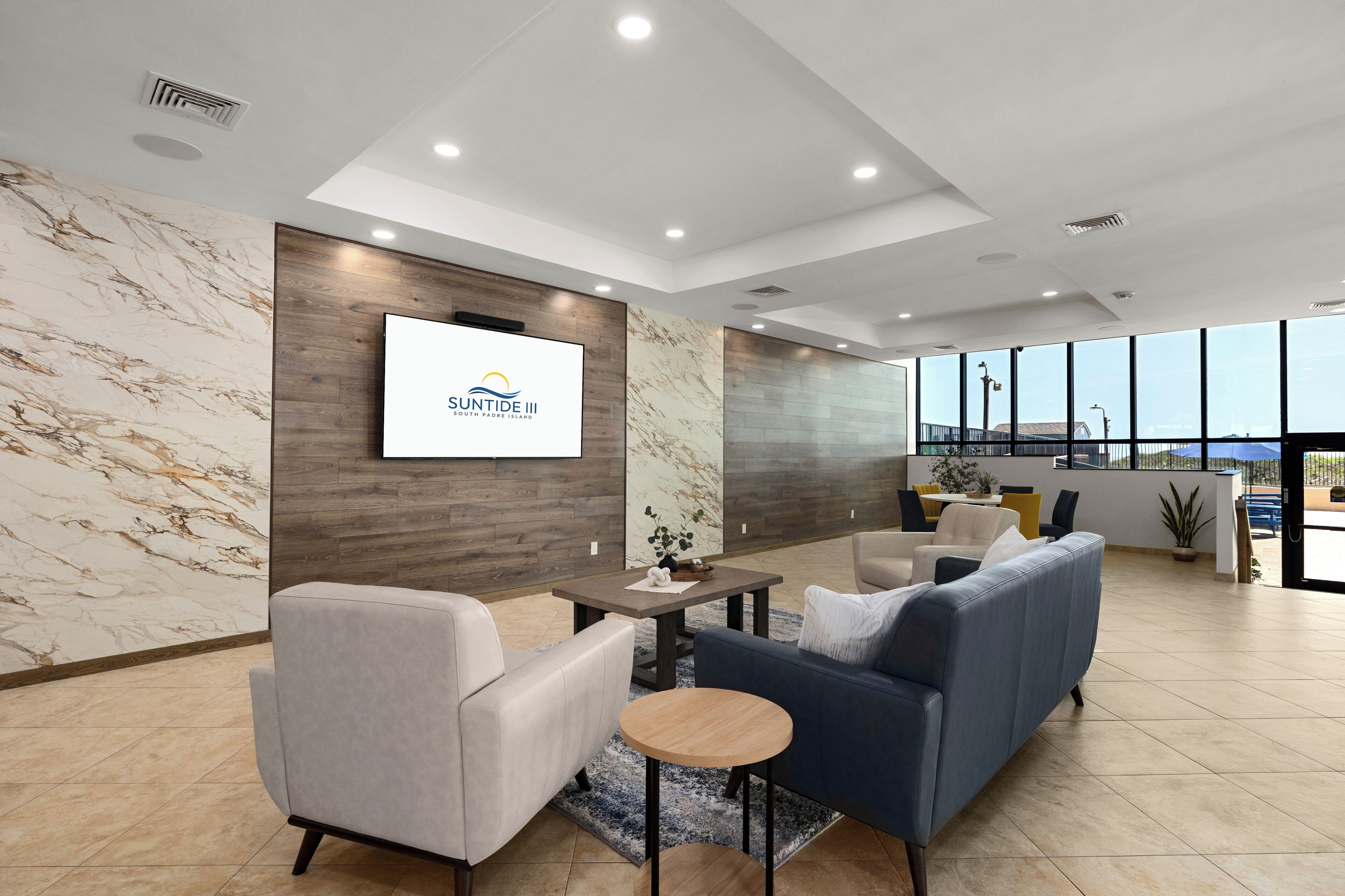 A modern hotel lobby with a marble wall, a mounted TV, a sofa, and a dining area with a view.