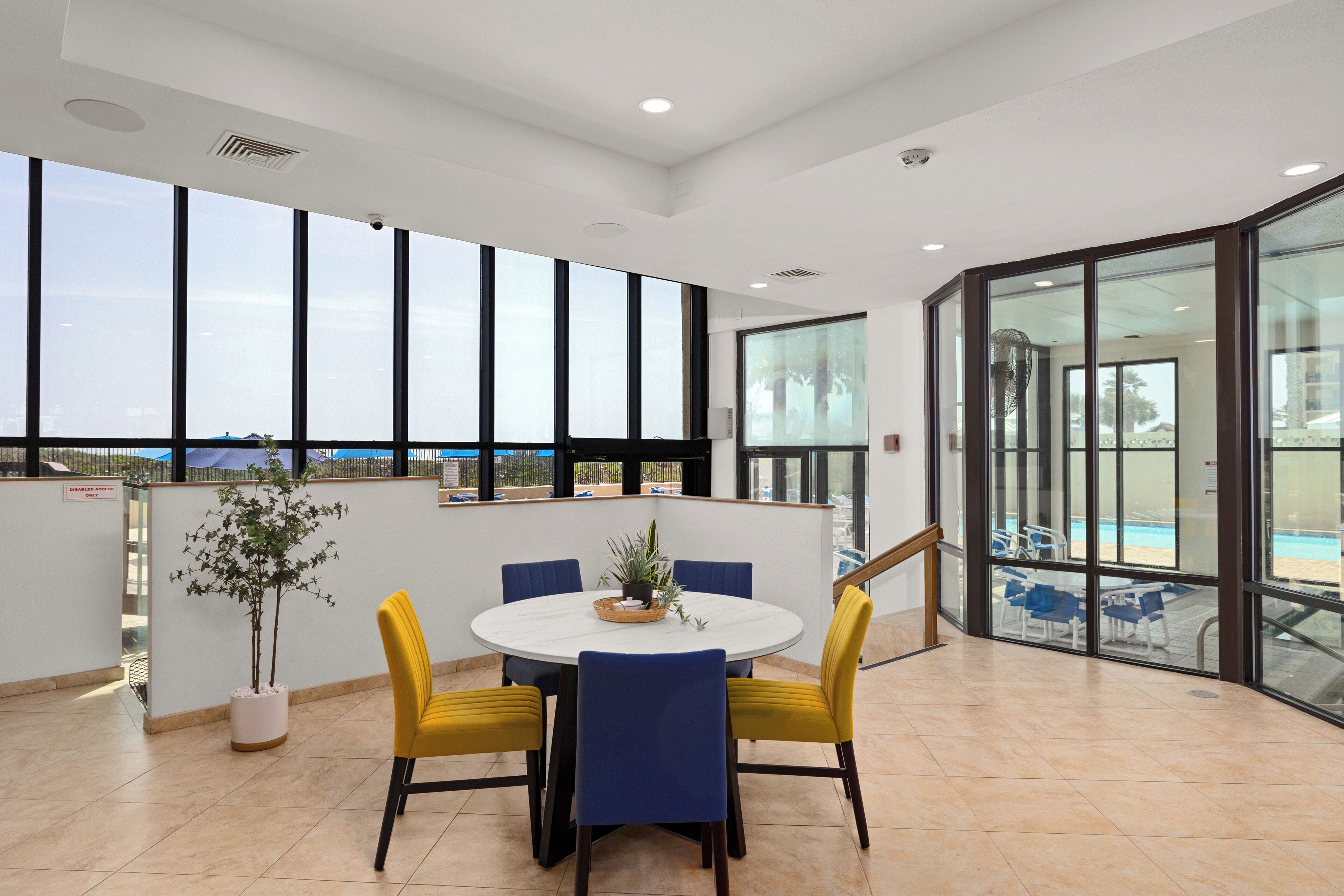 A spacious indoor area with a round table and chairs, large windows, and a view of a swimming pool.