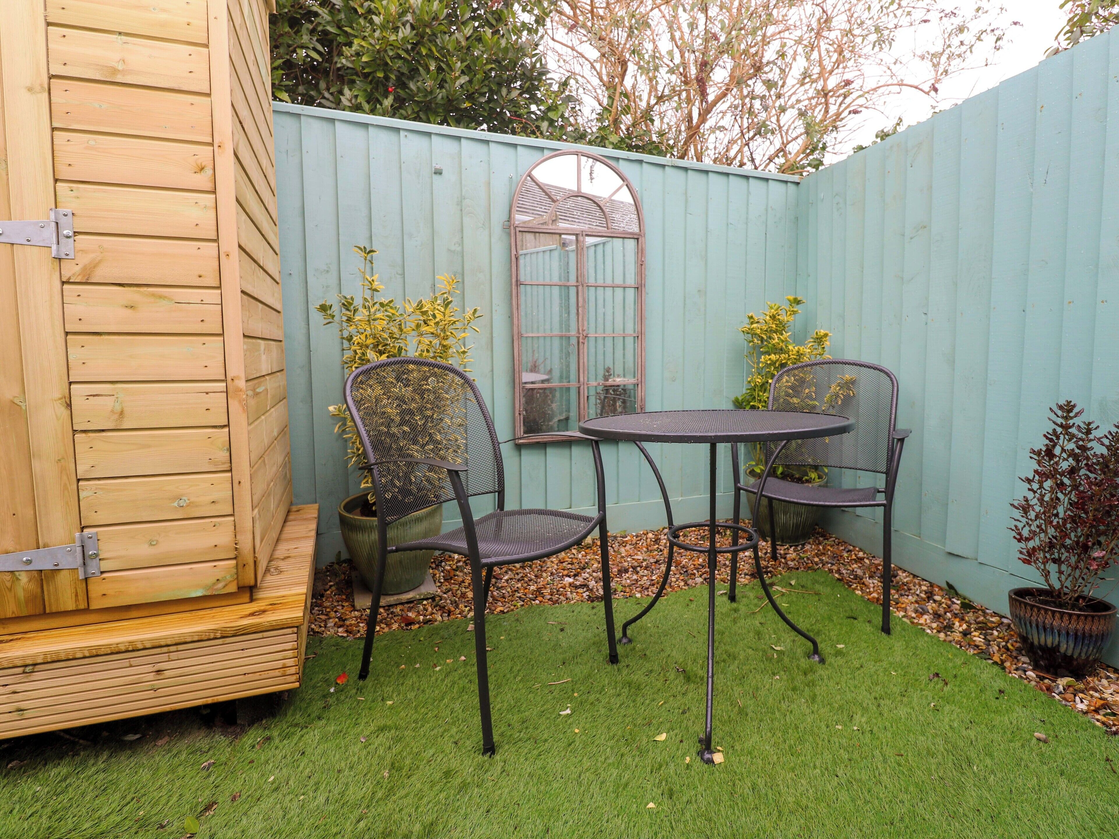 A small outdoor patio with a wooden deck, two metal chairs, a round table, and a potted plant.