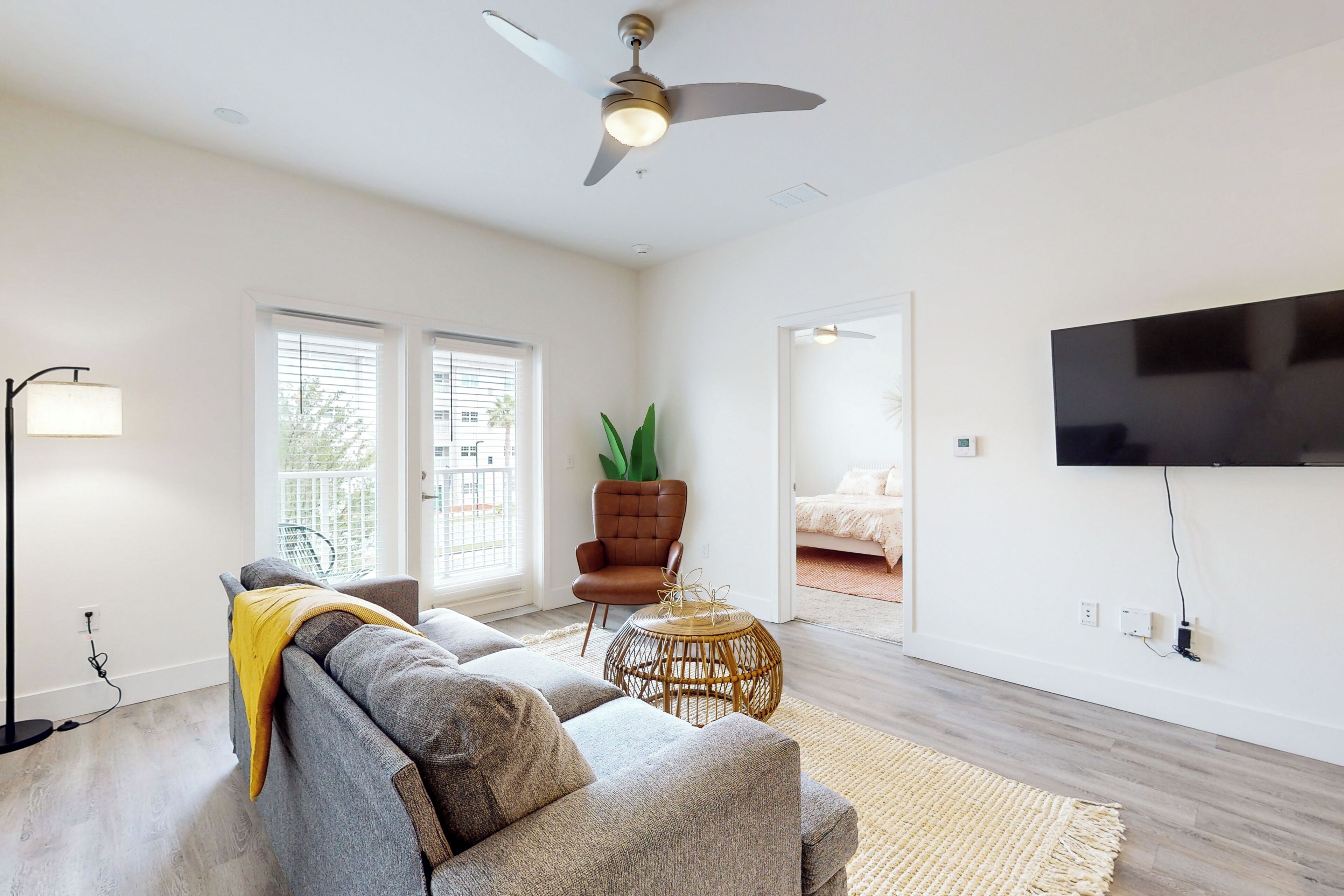 Un salon moderne avec un canapé gris, un fauteuil marron, une petite table d’appoint ronde en osier, un ventilateur de plafond et un téléviseur à écran plat fixé au mur.