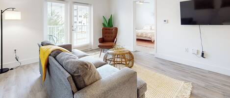 Un salon moderne avec un canapé gris, un fauteuil marron, une petite table d’appoint ronde en osier, un ventilateur de plafond et un téléviseur à écran plat fixé au mur.