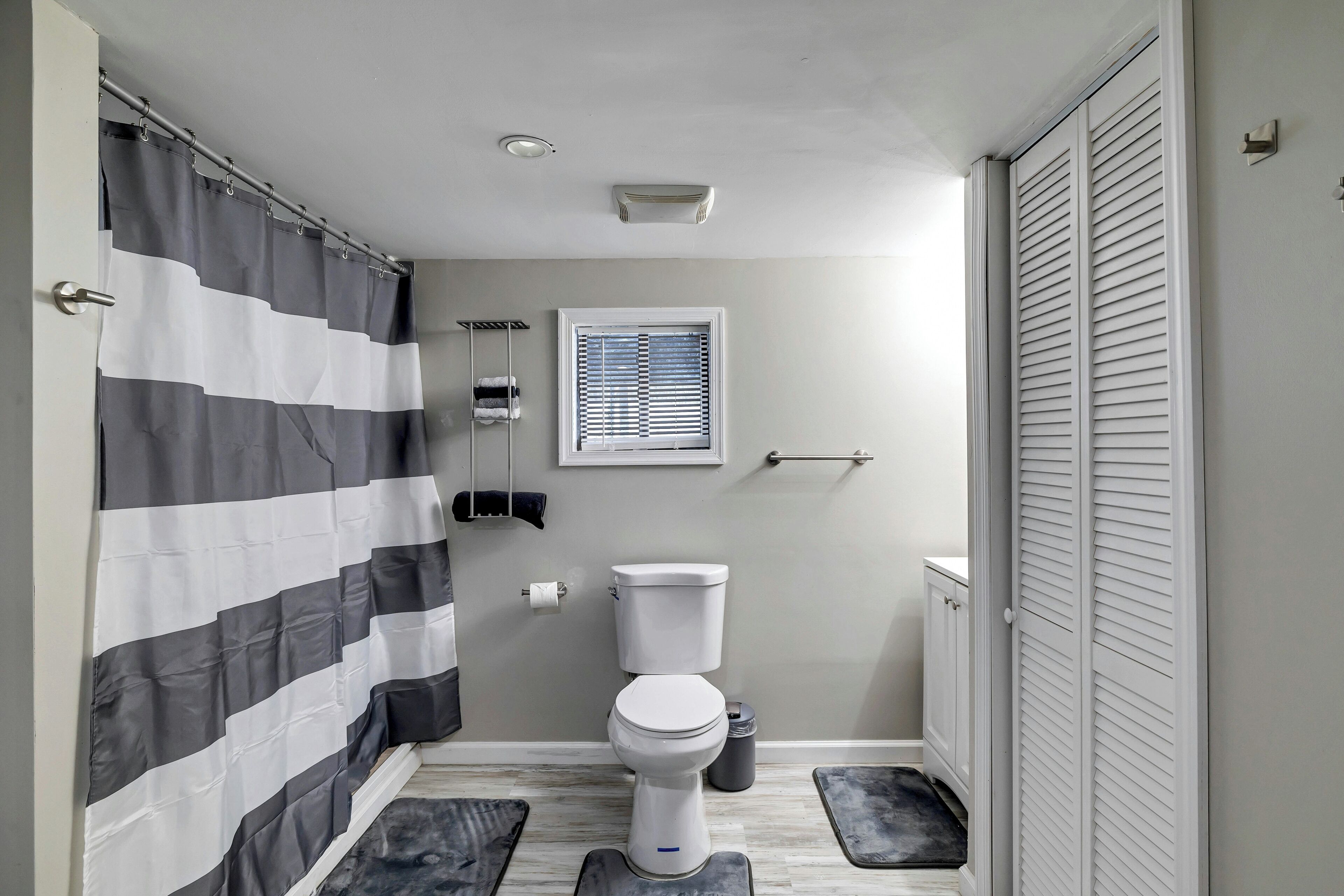 Un baño con cortina de ducha a rayas, un inodoro, una ventana con persianas y una puerta blanca con paneles verticales.