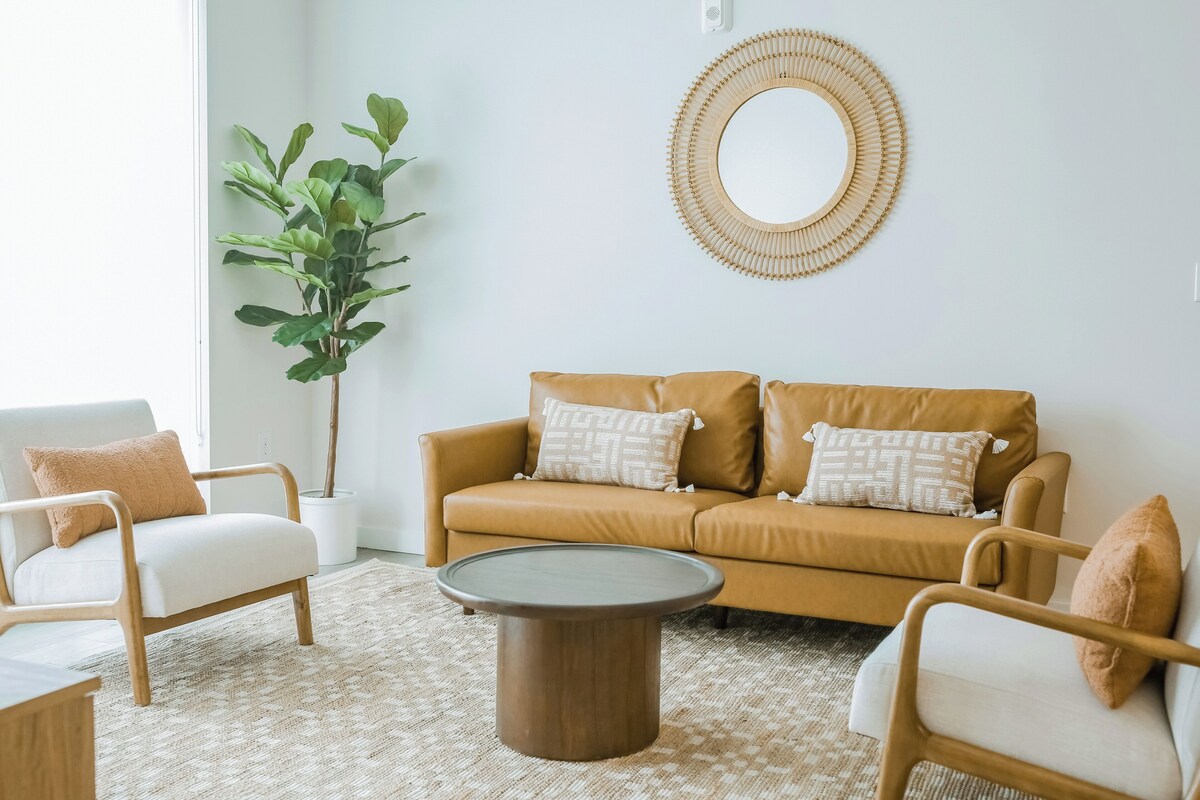 Un salón con un sofá color beige, dos sillones blancos, una mesa de centro redonda, una planta grande en maceta y un espejo montado en la pared.