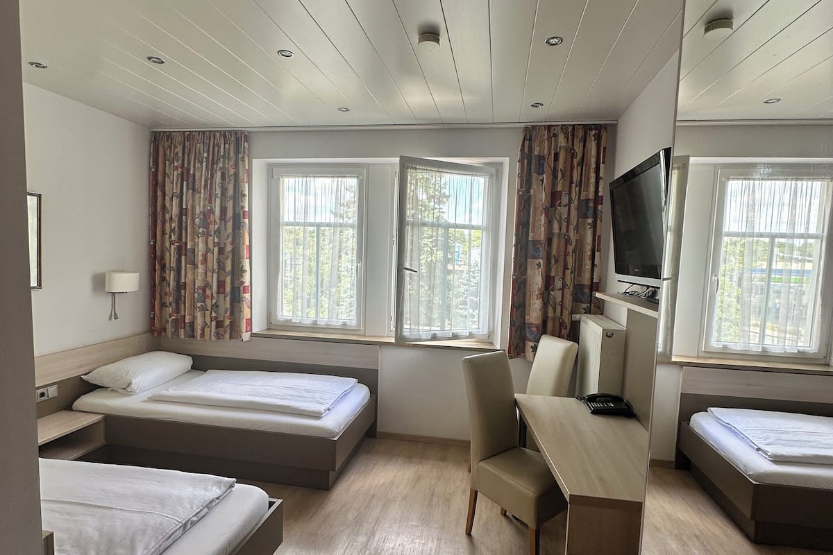 Habitación de hotel con tres camas, un escritorio, una silla, un televisor y ventanas grandes.