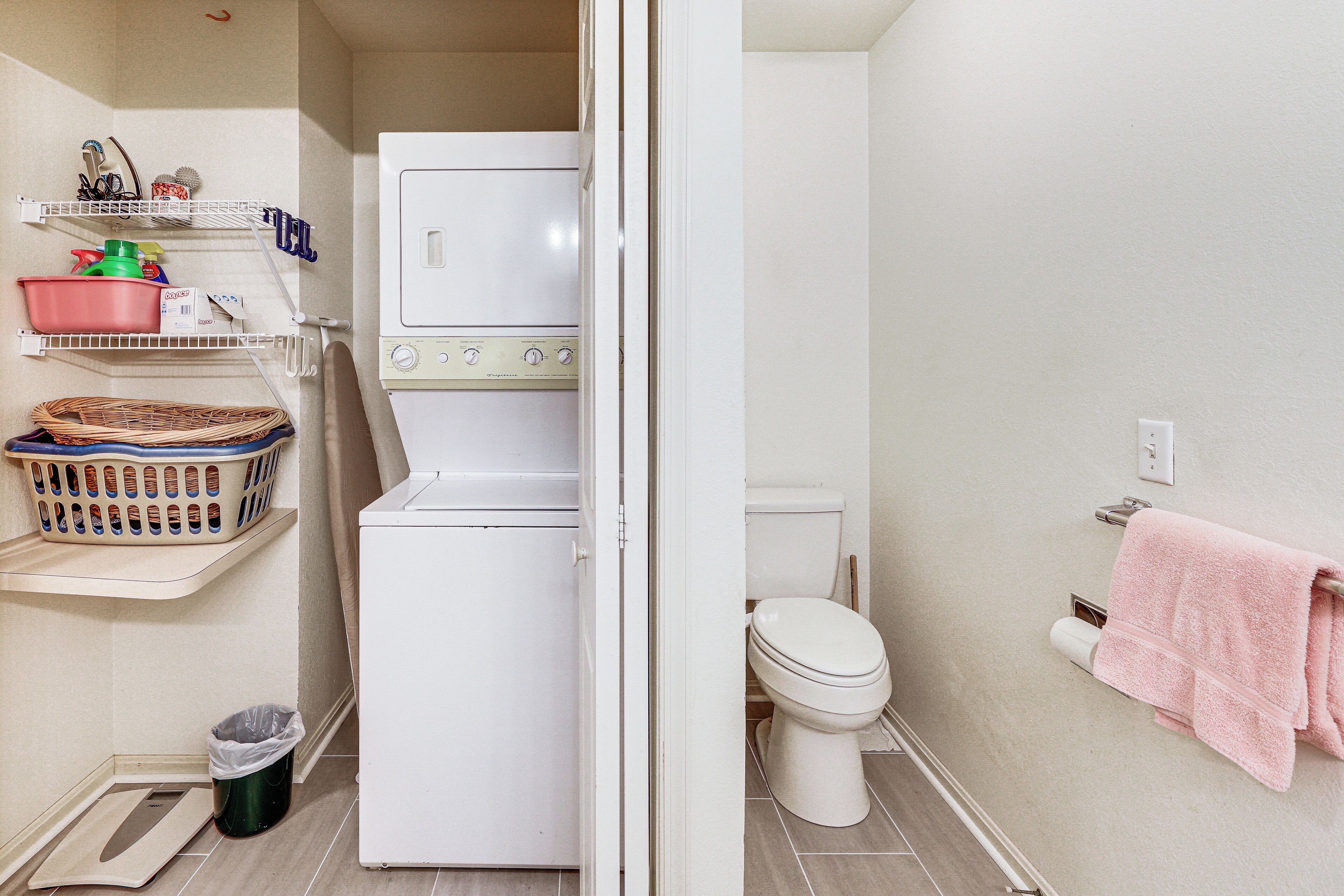 A compact bathroom with a washing machine, a toilet, a shelf with laundry baskets, and a trash bin.