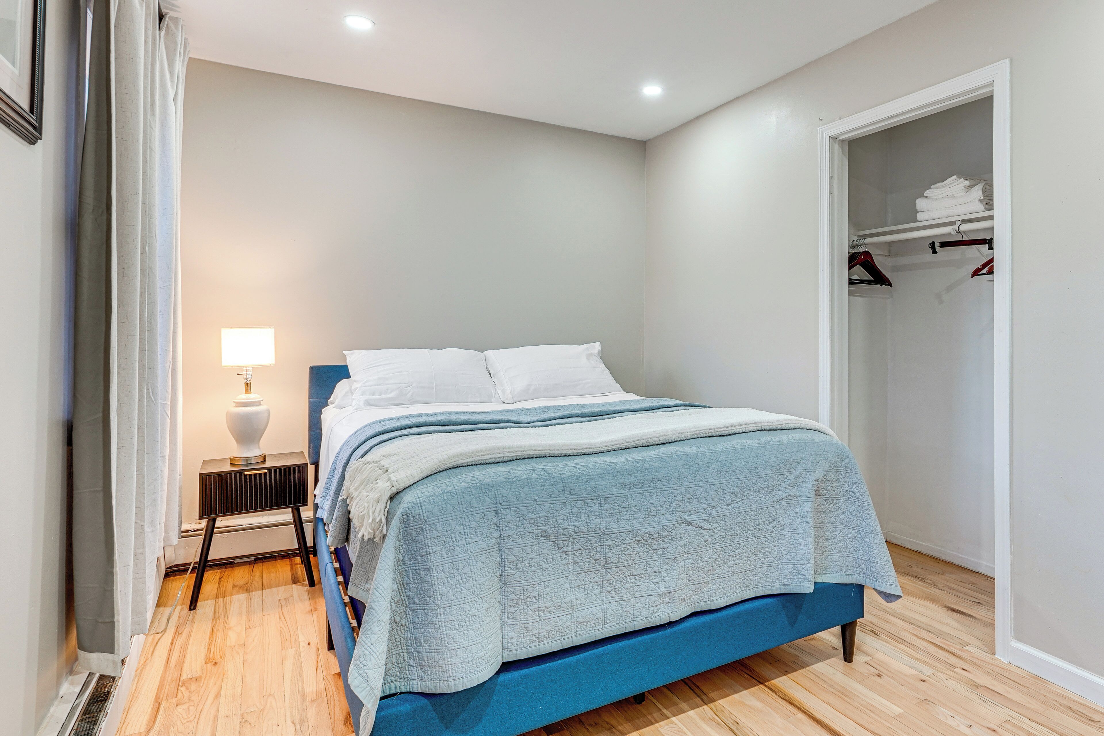 A bedroom with a bed, a bedside table with a lamp, a wooden floor, and a closet with hanging clothes.