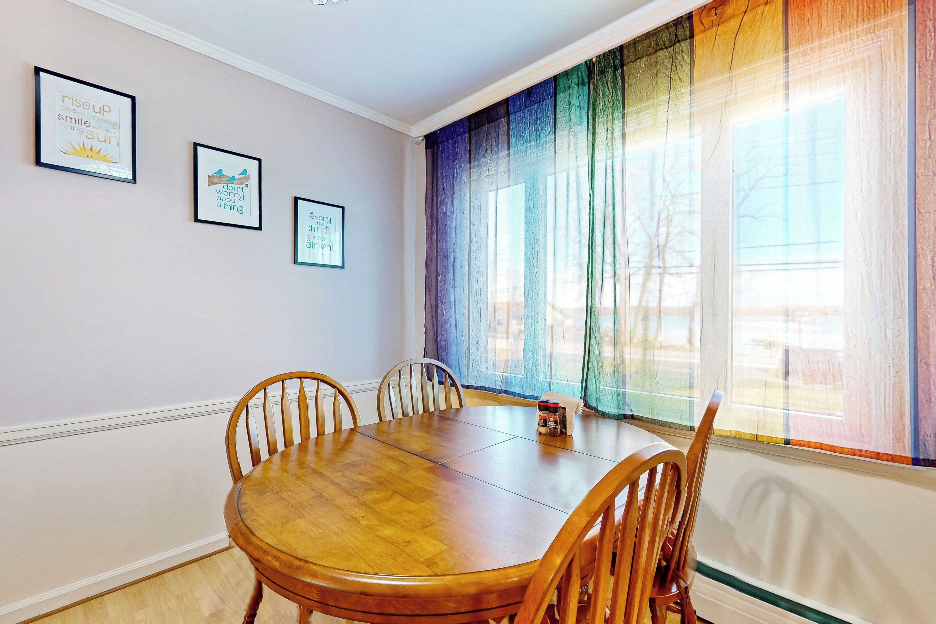 A dining area with a wooden table and chairs, framed motivational quotes on the wall, and a large window with colorful curtains.