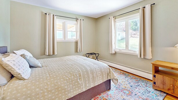 A bedroom with a bed, two windows with curtains, a wooden nightstand, and a colorful rug. - Lakefront 4BR w/ Stunning Views, Dock & Porch (Auburn)