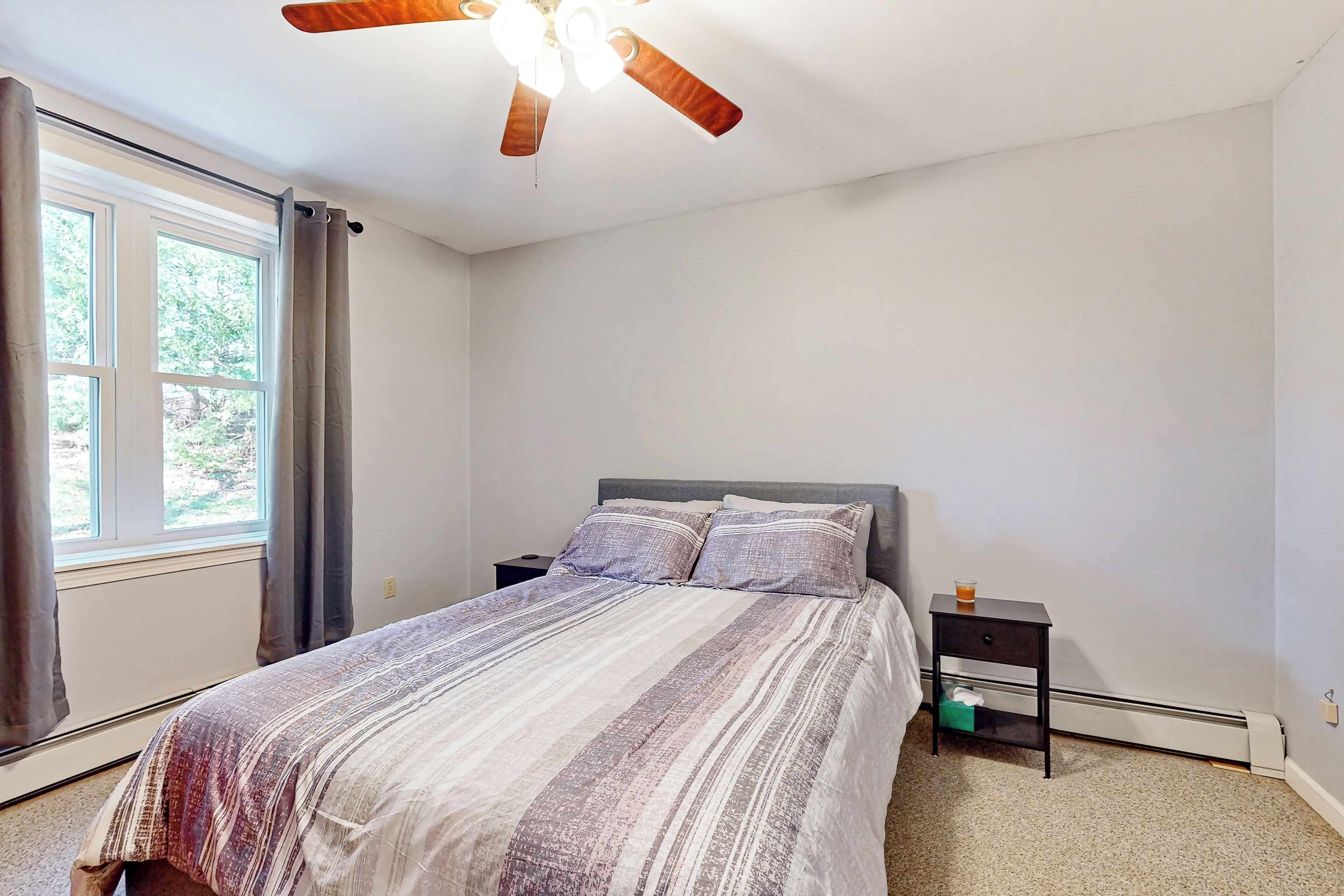 Un dormitorio con cama, mesita de noche, ventilador de techo, ventana con cortinas y radiador.