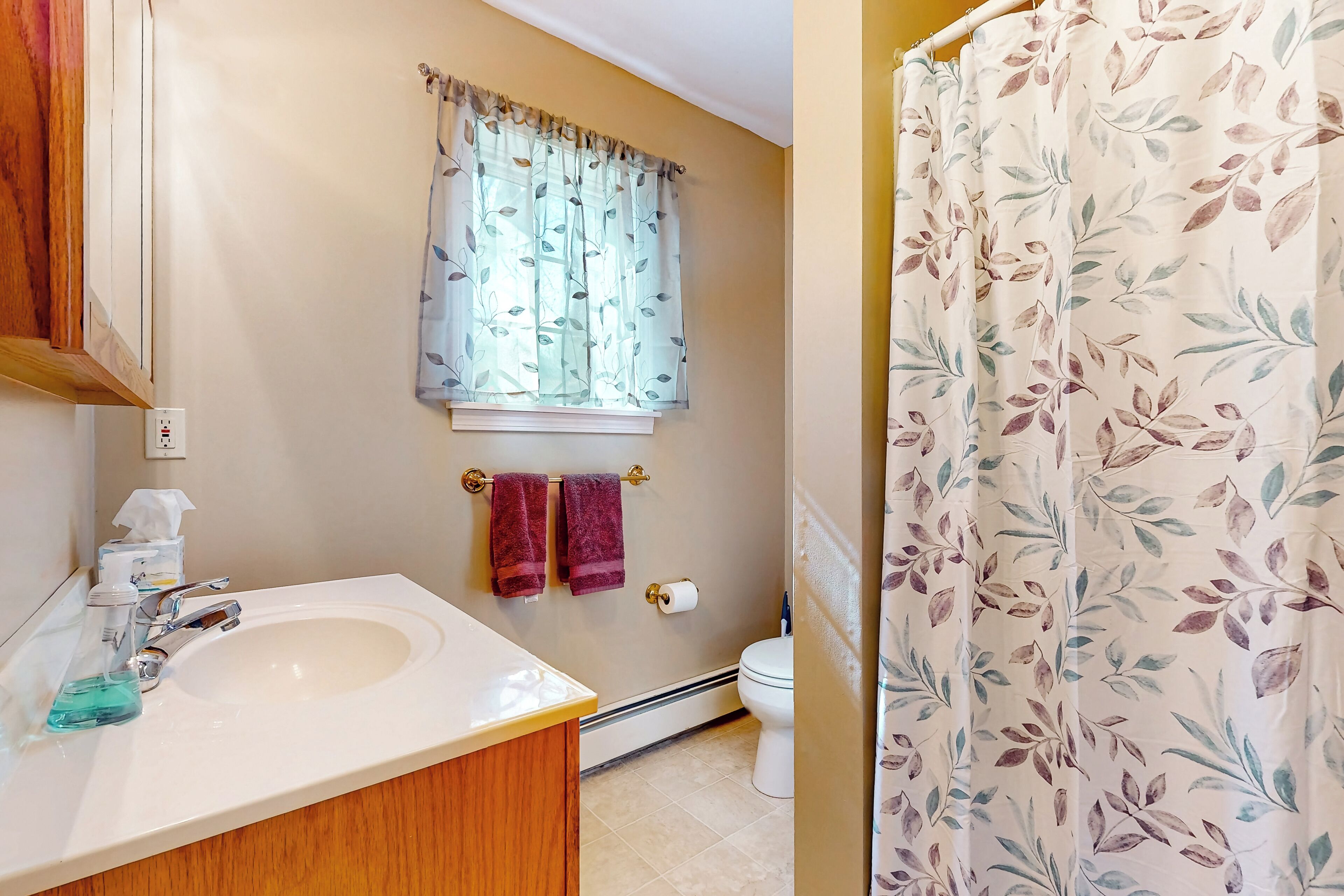 A bathroom with a sink, faucet, and a window with floral curtains. There are two towels hanging on a towel bar.