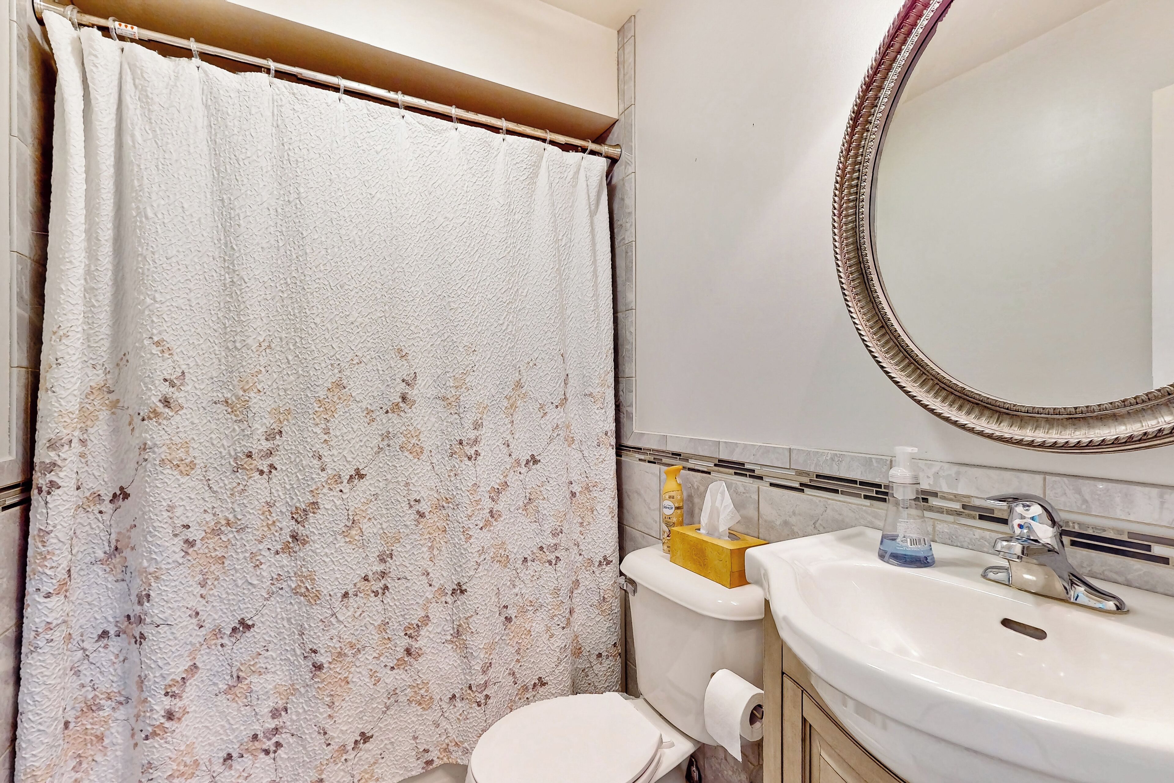 A bathroom with a floral shower curtain, a toilet, a sink with a mirror, and toiletries.