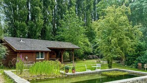 Un rustico chalet con uno stagno, immerso nel verde e circondato da alberi alti.