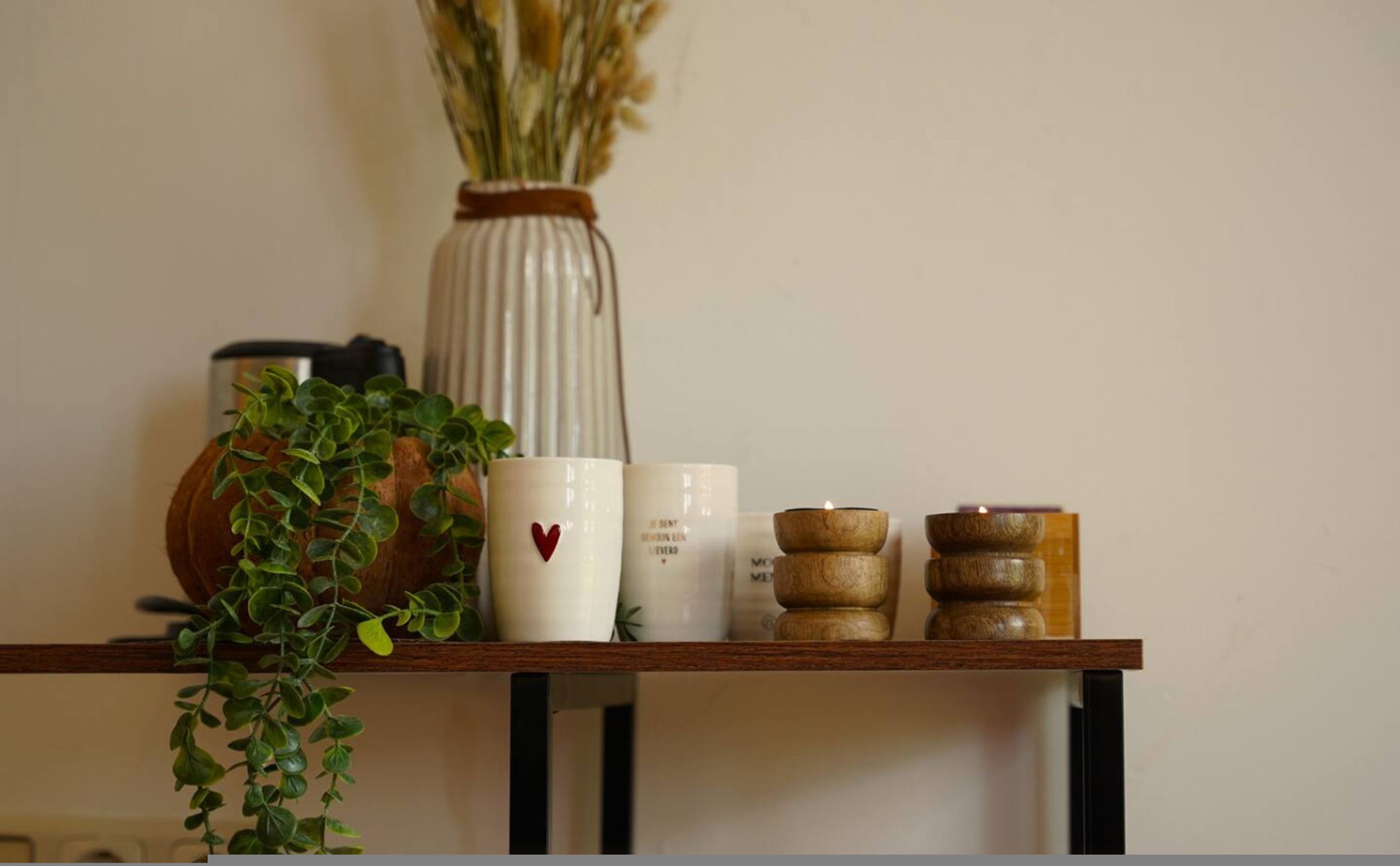 Uno scaffale con un vaso di fiori secchi, una pianta in vaso e diverse tazze bianche con scritte e un motivo a cuore rosso.