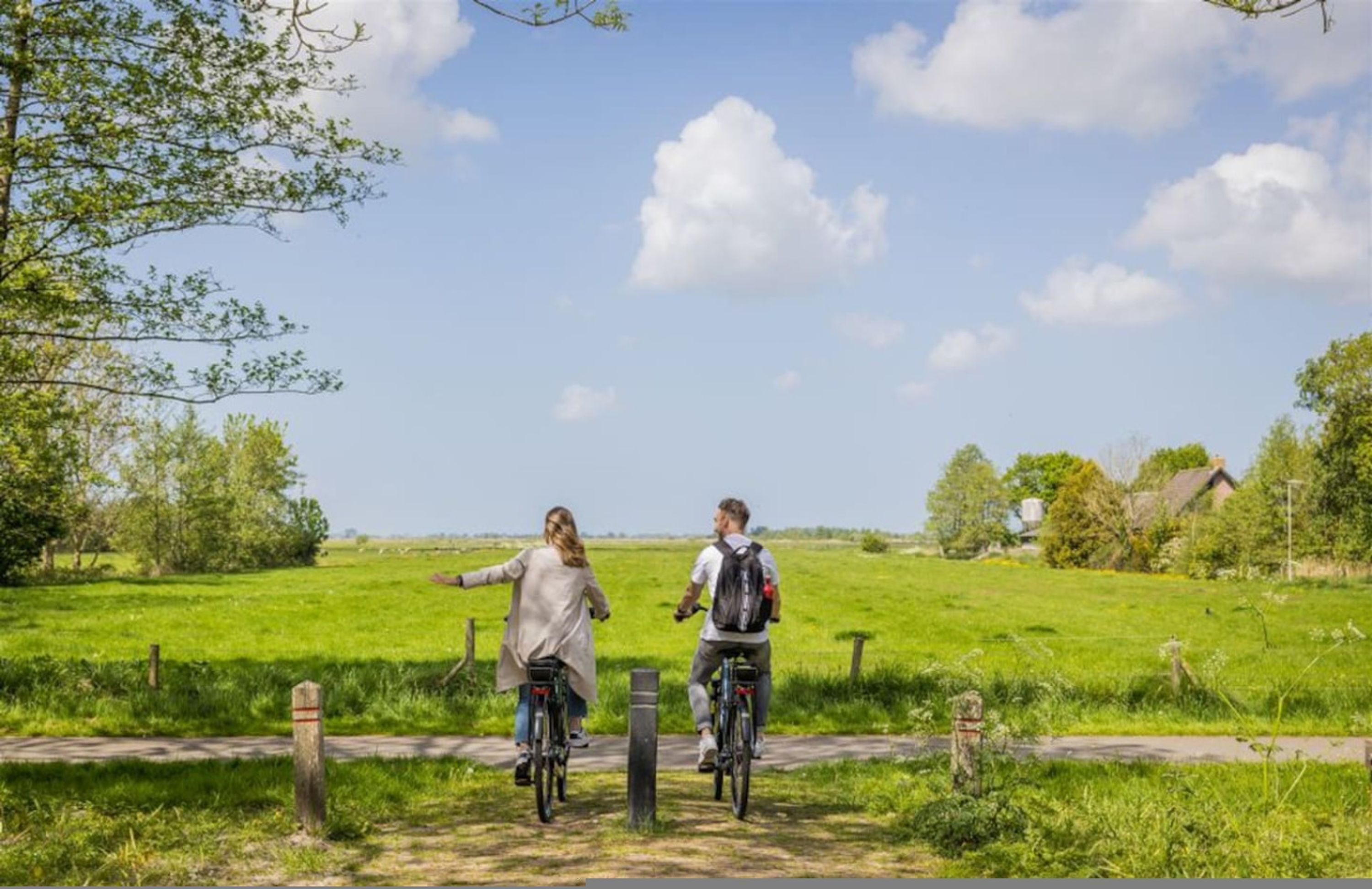Two people riding bicycles on a path in a rural area with green fields and a clear sky.