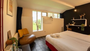 A modern hotel room with a large bed, a seating area with a brown chair, a small table with a vase, and a window with a view of greenery. - Biggemar Boerderijkamer (Weidum)