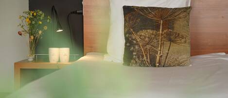 A neatly made bed with a white pillow and a decorative pillow with a nature-themed design. A bedside table with a lamp and a vase of flowers is visible.
