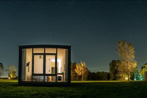 Estructura moderna con paredes de vidrio, ventilador de techo, cama y escritorio, sobre un cielo nocturno estrellado.