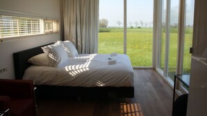 A bedroom with a large bed, a view of a grassy field through large windows, and a red chair. - Anjewiel Weidesuite in Weidum (Weidum)