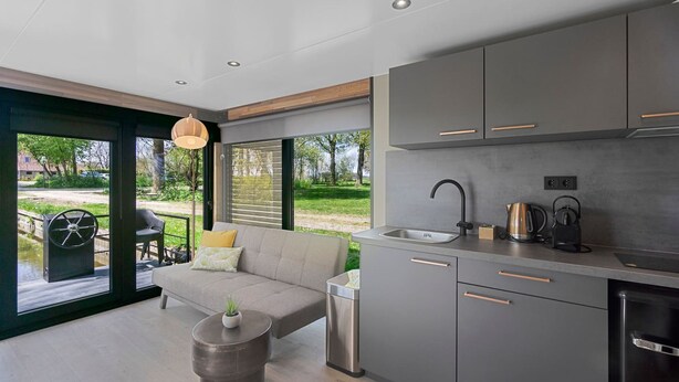 A modern kitchen with grey cabinets, a stainless steel sink, and a kettle. There is a sofa and a small table with a plant. - Doeke Sleat Houseboat (Weidum)