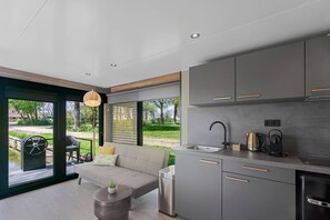 A modern kitchen with grey cabinets, a stainless steel sink, and a kettle. There is a sofa and a small table with a plant. - Doeke Sleat Houseboat (Weidum)