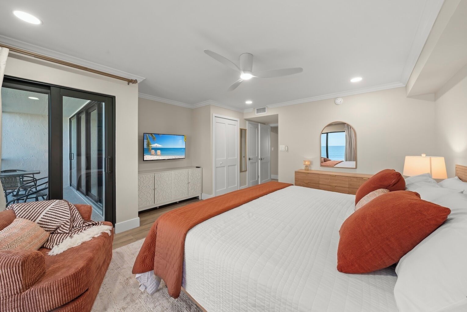 A bedroom with a large bed, a ceiling fan, a TV on the wall, and a sliding glass door leading to a balcony.