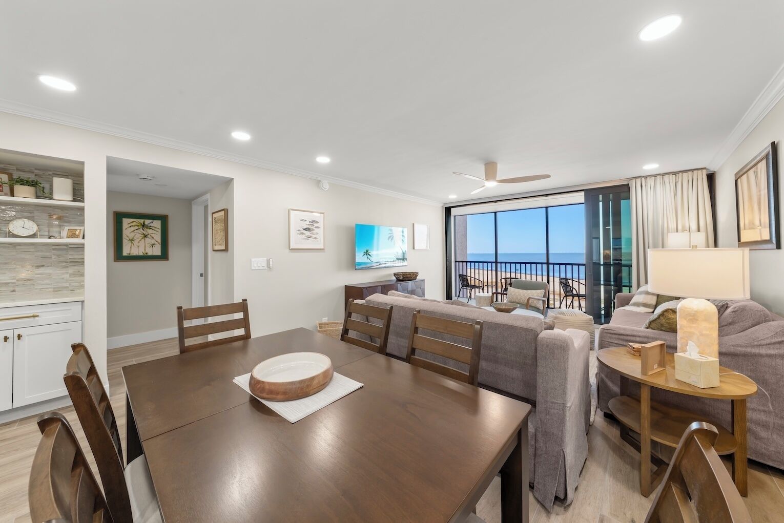 A modern living room with a dining area, large windows with a view of the sea, and a balcony with outdoor seating.
