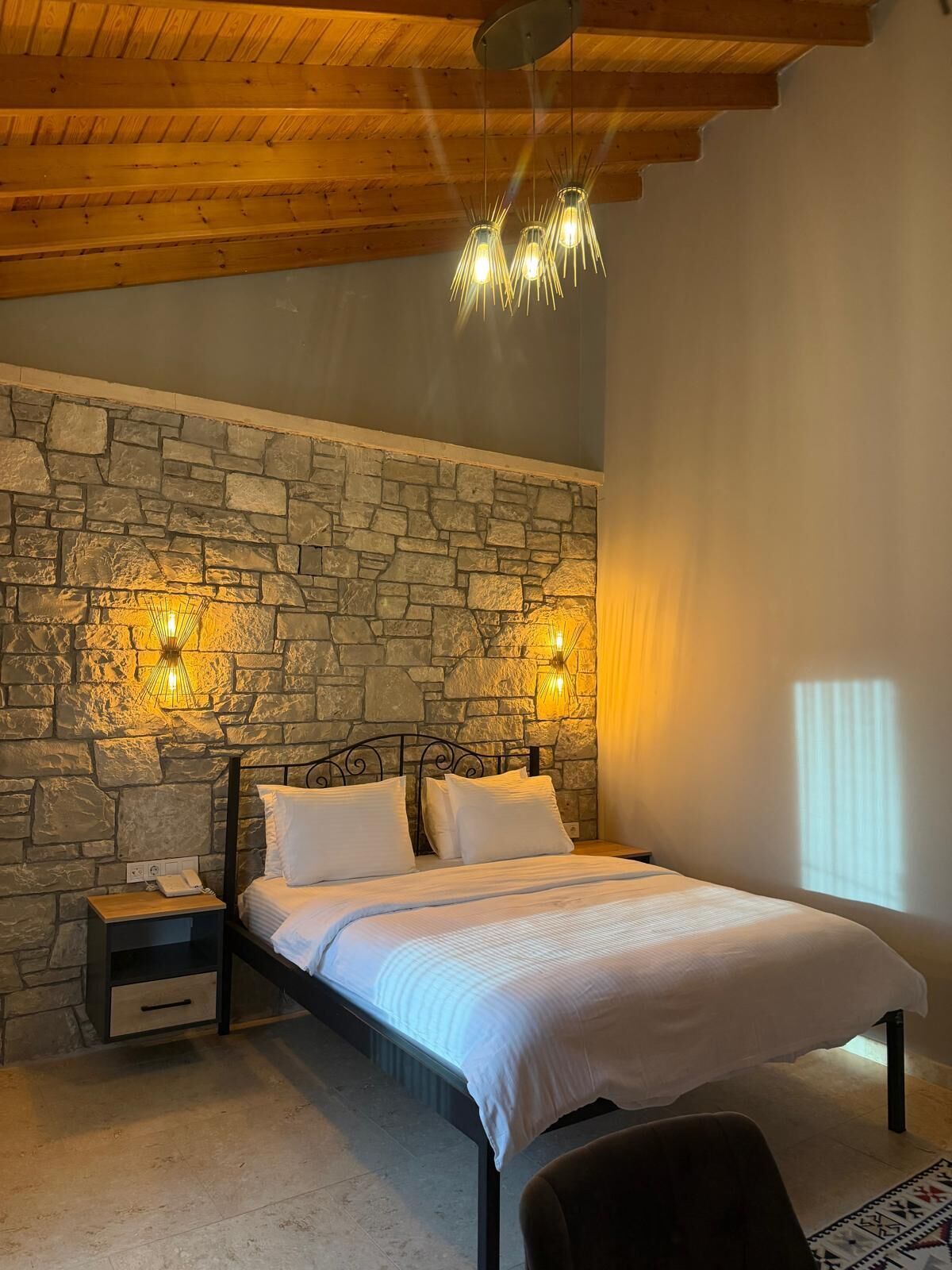 Un dormitorio con pared de piedra, techo de madera, cama con ropa blanca y dos lámparas de noche.