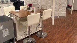 A dining area with a wooden table and four white chairs, a kitchen area with a stove and refrigerator, and a white partition with a patterned design.