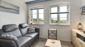 Une pièce avec un canapé en cuir, une petite table en bois, une lampe et une vue sur la campagne par deux fenêtres.