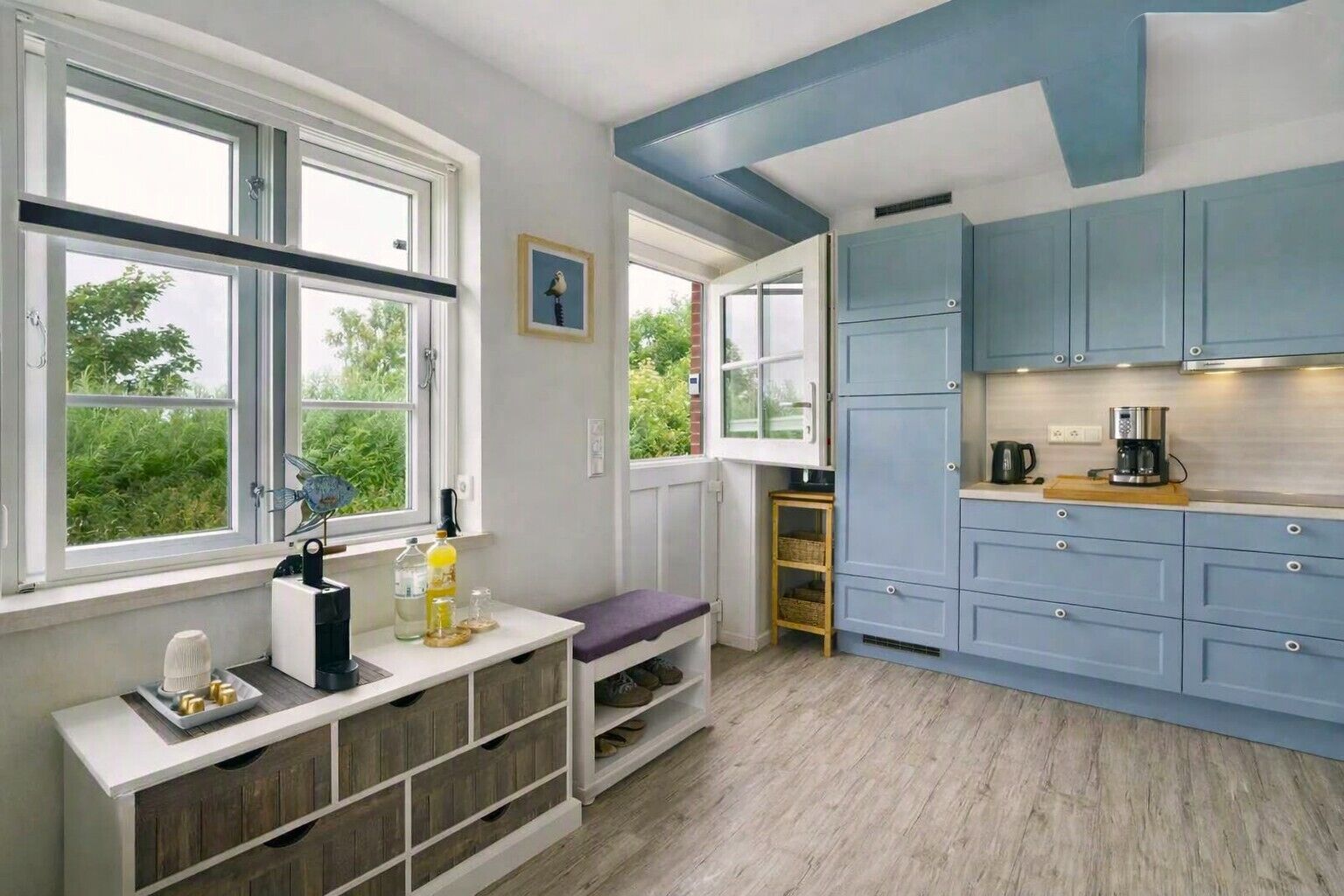 A modern kitchen with light blue cabinets, a wooden bench, and a view of greenery outside.