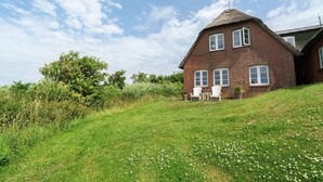 Une maison en briques avec un toit de chaume, des fenêtres blanches et une cour verdoyante où se trouve une chaise.