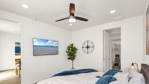 Un dormitorio con cama, ventilador de techo, televisión instalada en la pared, una planta y una puerta que da a otra habitación.