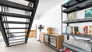Quarto moderno com escada de metal, armário de madeira com gavetas, estante e um pequeno ventilador.