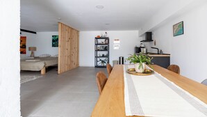 Amplo espaço de estar com uma mesa de jantar de madeira, cozinha ao fundo e uma cama no canto.