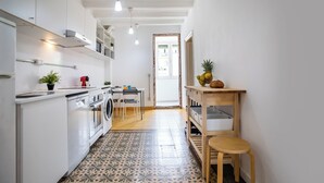 A narrow kitchen with white cabinets, a washing machine, a dining area with a table and chairs, and a wooden stool. - BBarcelona Sants Flat (Barcelona)
