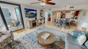 A spacious living room with a ceiling fan, a television, a dining area with a kitchen in the background, and a large window. - Mainsail 321 2 Bedroom Condo (Miramar Beach)