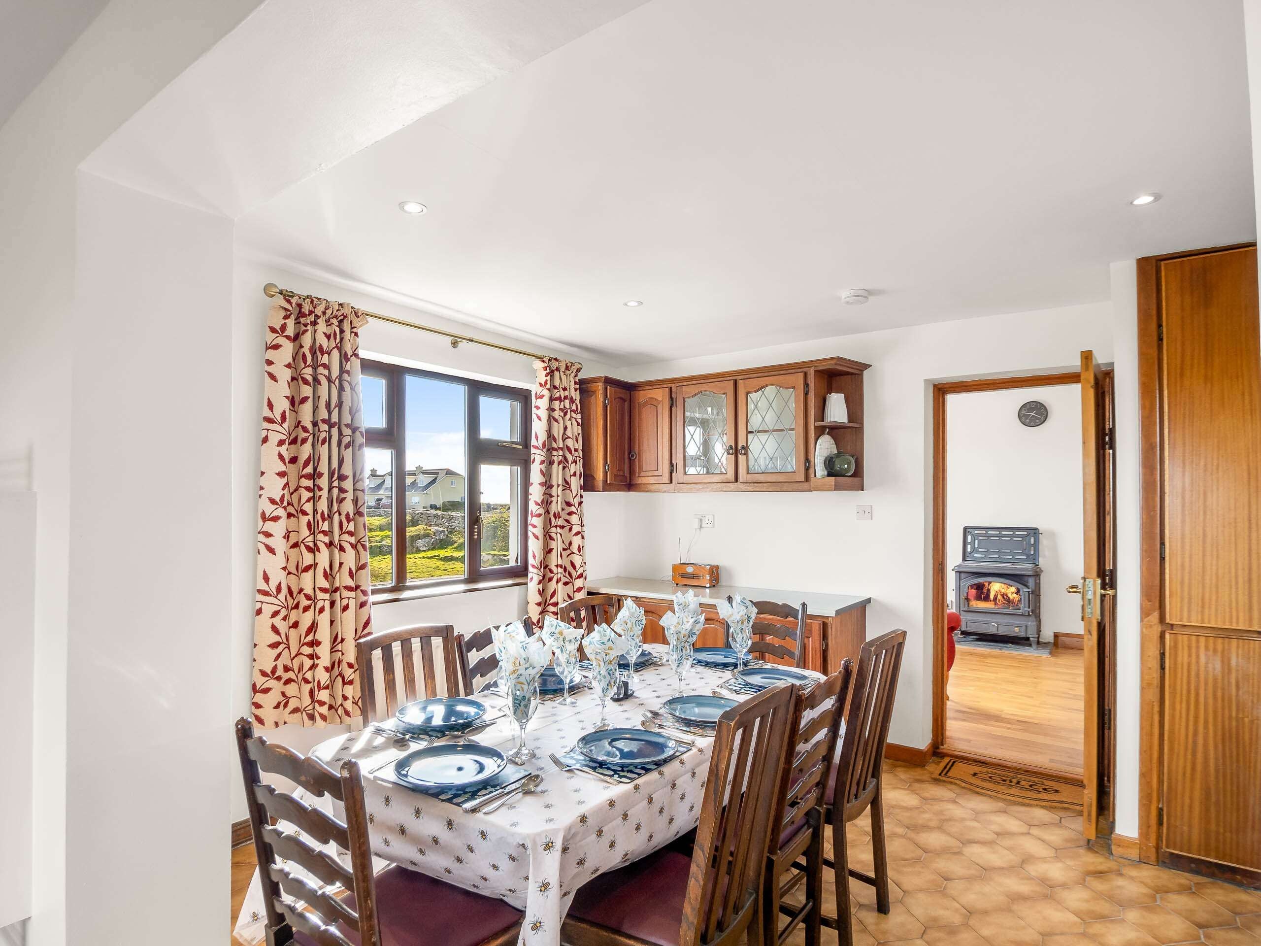 A dining room with a table set for four, wooden chairs, a window with curtains, and a kitchen area with wooden cabinets and a stove.