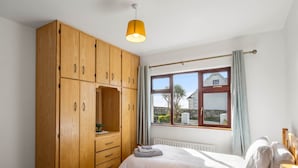 Ein Schlafzimmer mit einem Holzkleiderschrank, einem Bett mit weißer Bettwäsche, einem Nachttisch mit Lampe und einem Fenster mit Vorhängen.