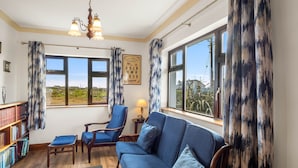Ein gemütliches Wohnzimmer mit einem blauen Sofa, Holzsesseln, einem Bücherregal und großen Fenstern mit Vorhängen.
