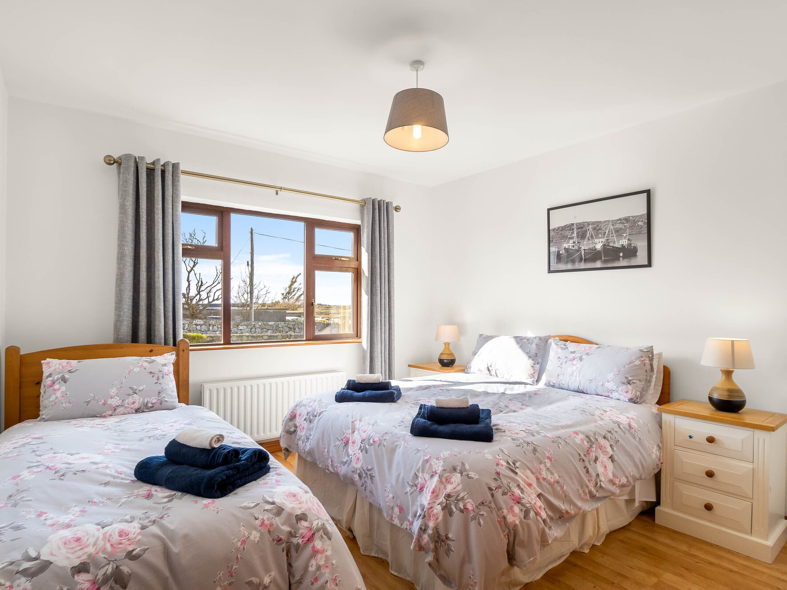 A bedroom with two beds, a window with curtains, a bedside table with a lamp, and a framed picture on the wall.
