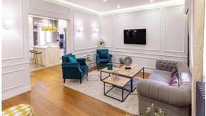 Een moderne woonkamer met een grijze bank, smaragdgroene fauteuils en een salontafel. Een flatscreen televisie is aan de muur bevestigd, en er is een keuken te zien op de achtergrond.